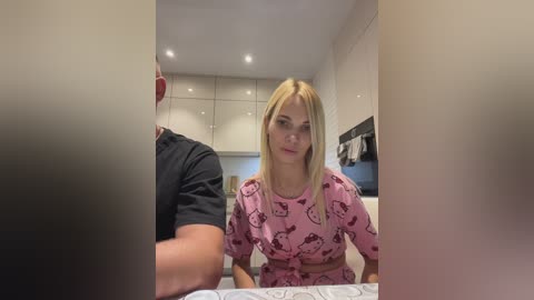 Media: Video of a young blonde woman in pink Hello Kitty pajamas, sitting in a modern kitchen with white cabinets, recessed lighting, and a black shirt-wearing man partially visible.