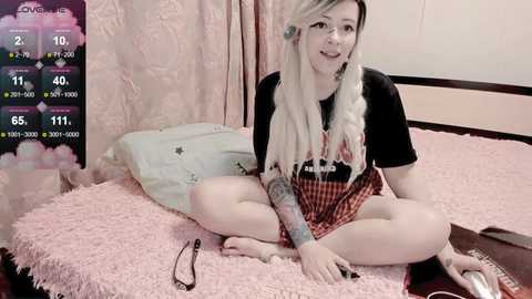 A video of a blonde, tattooed woman with long hair in a black t-shirt and red plaid skirt, sitting cross-legged on a pink bed with a TV displaying a video game.