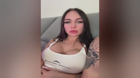Media: Video of a curvy woman with long black hair, wearing a white crop top revealing ample cleavage, sitting on a gray couch, with a skull tattoo on her left arm.