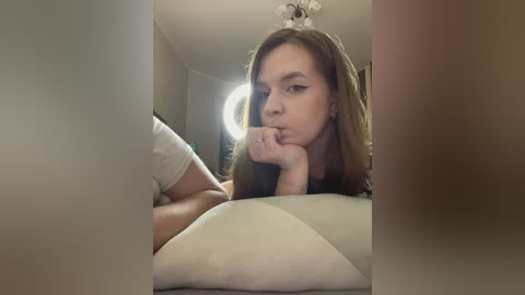 Media: Video of a young woman with long brown hair, wearing a white shirt, resting her chin on her hand, gazing introspectively. The background features a beige wall and a round mirror, creating a soft, intimate atmosphere.