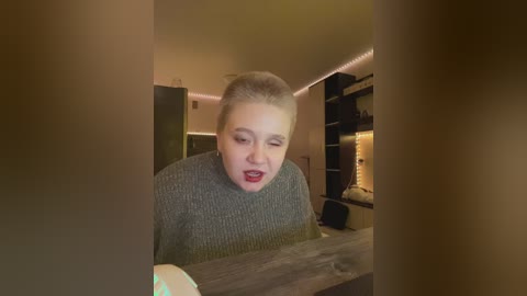 A candid video of a woman with light skin and blonde hair, wearing a grey sweater, looking surprised while holding a white plate. The background features a modern kitchen with wooden cabinets and a countertop.