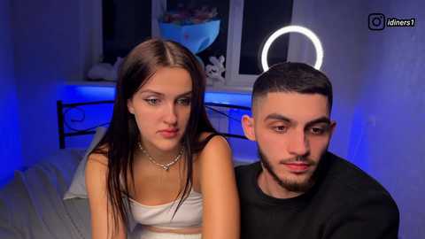 Video of a young woman with long brown hair in a white crop top and a young man with short dark hair in a black shirt, sitting in a room with blue lighting, a neon halo, and a bed in the background.