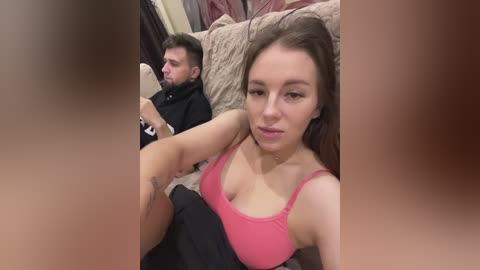 Media: A candid video captures a woman with light skin, brown hair, and a pink top, sitting on a beige couch, holding a phone. Behind her, a bearded man in a black hoodie is seated, looking away.