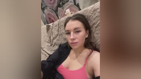 Media: A video of a young woman with fair skin, brown hair, and wearing a pink tank top, lying on a beige quilted bedspread. The background features floral wallpaper.