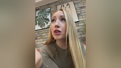 Media: Video of a fair-skinned, blonde woman with long hair, wearing a gray top, looking contemplative, in a modern, brick-walled room with green leaf art.