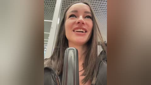 Media: Video of a smiling Caucasian woman with straight brown hair, wearing a gray jacket, standing near a gray microphone inside a modern office with white ceiling tiles.