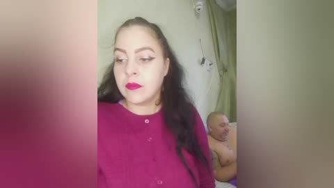 Video of a young woman with light skin, long black hair, and red lipstick, wearing a purple top, standing in a room with a baby in a white crib.