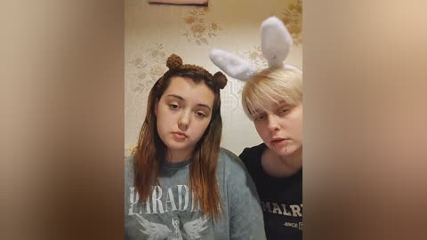 Media: Video of two young women wearing bunny ears, one with brown hair and the other with blonde hair, both in gray sweaters, indoors with floral wallpaper background.