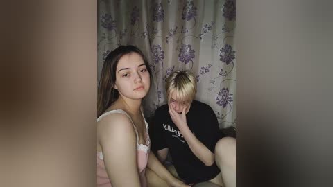 Media: Video of two young women, one with long dark hair in a pink tank top, the other with short blonde hair in a black t-shirt, both sitting, looking sad, in a room with floral-patterned curtains.