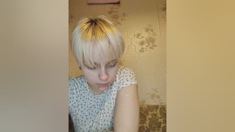 Media: Video of a young, fair-skinned, short-haired woman with blonde hair, wearing a white floral-patterned top, looking down, with a beige floral wallpaper background.