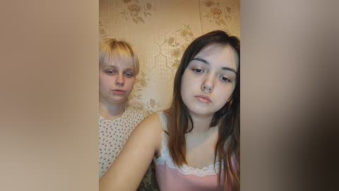 Media: Video of two young women with fair skin; one with blonde hair, one with brown hair, both wearing casual clothing, against a floral wallpaper background.