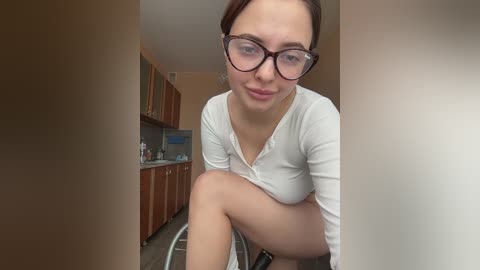 Media: A video of a fair-skinned woman with glasses and brown hair, wearing a white long-sleeve shirt, crouching in a modern kitchen with wooden cabinets and stainless steel appliances.