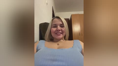 Media: Video of a smiling, light-skinned, blonde woman with shoulder-length hair, wearing a light blue ribbed tank top, standing in a room with wooden furniture and beige walls.