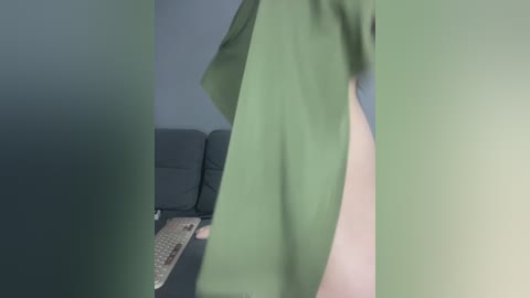 Media: A video of a person wearing a green shirt, standing in a dimly lit room with a dark grey couch and a beige remote control on the floor. The background is blurred.