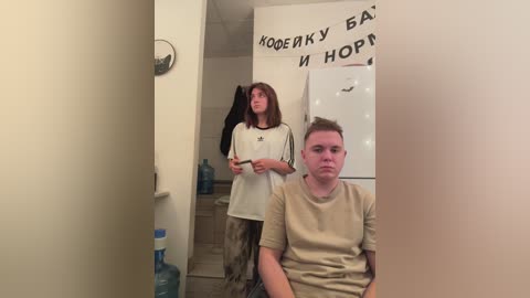 Media: A video of a young man with short brown hair, wearing a beige T-shirt, standing in front of a mirror. Behind him, a woman with long brown hair, wearing a white shirt, is reflected.