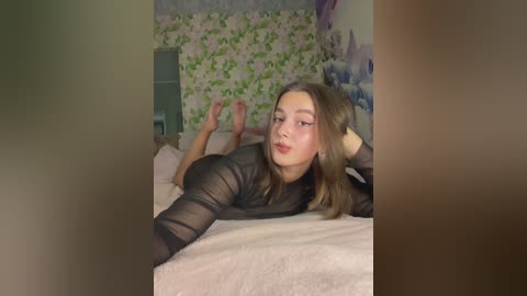 A video of a young Caucasian woman with long brown hair lying on a bed, wearing a sheer black top, in a softly lit bedroom with floral wallpaper and a mirror.