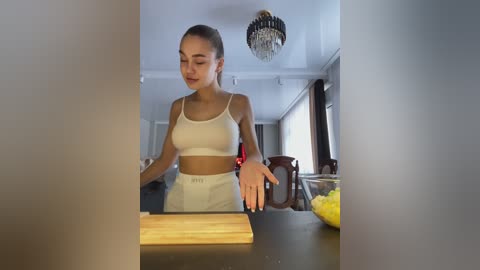 Media: Video of a fit young woman with light skin and brown hair tied back, wearing a white sports bra and high-waisted pants, chopping vegetables on a dark kitchen island. Background features a chandelier and large windows.