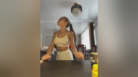 Media: Video of a slender, fit woman in a white sports bra and matching high-waisted leggings, mid-yoga stretch, in a modern living room with large windows and a chandelier.