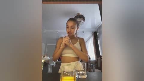 Media: A video of a young woman with light skin, long brown hair in a high bun, wearing a white crop top and high-waisted shorts, standing in a modern, well-lit living room with a wooden table, glass, and a plant in the foreground.