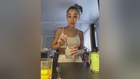 Media: Video of a young woman with dark hair in a bun, wearing a white sports bra and shorts, blending a smoothie in a kitchen with large windows.