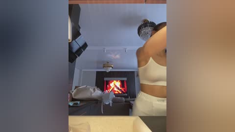 Media: A video of a woman in a white crop top and pants standing near a lit fireplace in a modern living room with a ceiling fan.