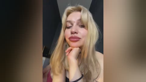 Video of a blonde woman with fair skin and light makeup, resting her chin on her hand, smiling slightly. She wears a black top. Background is blurred, showing a dark room with a white ceiling.