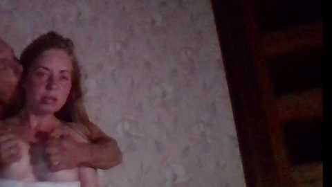 Video depicting a nude, fair-skinned woman with long, blonde hair, her face in distress, held by a muscular, dark-haired man with a tattooed arm. The setting is dimly lit, with a rustic wooden background.