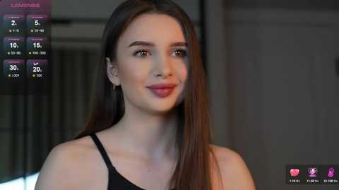 Media: Video of a young woman with long, straight brown hair, fair skin, and a slim physique. She wears a black tank top and smiles softly. Background shows a blurred indoor setting.