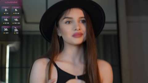 Media: Video of a young woman with long brown hair, wearing a black hat and sleeveless top, indoors against a blurred background.