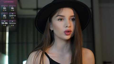 Media: Video of a young woman with long brown hair, wearing a black hat and black top, standing indoors. Background is blurred, showing a modern interior.