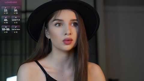 Media: Video of a young woman with fair skin, long brown hair, wearing a black hat and black top, looking introspective in a dimly lit room.