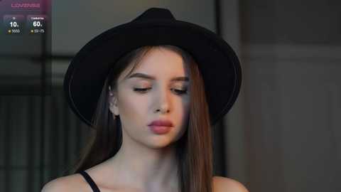 Media: Video of a young woman with fair skin, long straight brown hair, and a black wide-brimmed hat. She wears a black top, has a serene expression, and stands indoors with blurred, muted background.