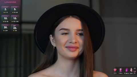 Media: Video of a young woman with light skin, long brown hair, and a black hat, smiling in an indoor setting with a dimly lit background.