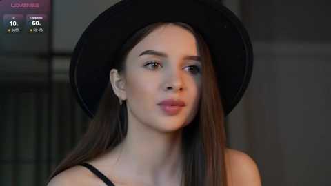Media: Video of a young woman with fair skin and long, straight brown hair, wearing a black hat and black top. Background is blurred, showing an urban setting.