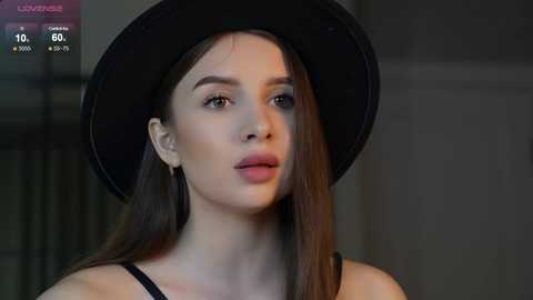 Media: Video of a fair-skinned woman with long brown hair, wearing a black hat and black top, looking contemplative against a blurred background.