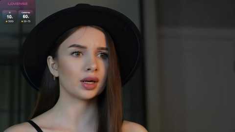Media: Video of a young woman with fair skin, straight brown hair, and a black hat, wearing a black top, against a blurred, dimly-lit background.