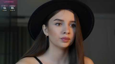 Media: Video of a young woman with fair skin, long straight brown hair, and wearing a black hat and black top, indoors with blurred background.
