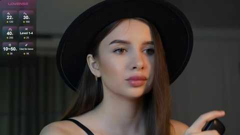 Media: Video of a young woman with straight brown hair, wearing a black hat and a black top, with a blurred indoor background.