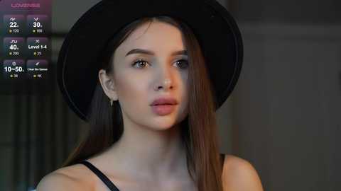 Media: Video of a young woman with fair skin, long brown hair, wearing a black hat and black top, in a dimly lit room with a video game overlay.