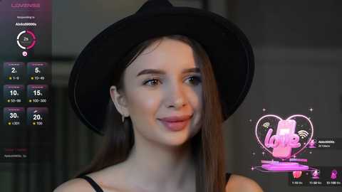 Media: Video of a young woman with fair skin, long dark hair, and a black hat, smiling, against a blurred, dimly lit background. She wears a black top.