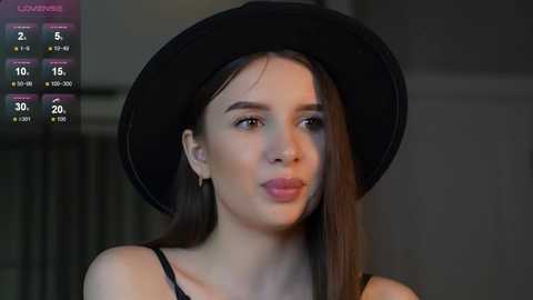 Media: Video of a fair-skinned woman with straight brown hair, wearing a black hat and black top, standing against a dimly lit, blurred background.