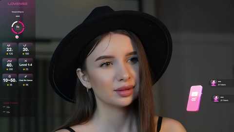 Media: Video of a young woman with fair skin, long brown hair, and a black hat, wearing a black top, standing indoors.