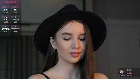 Media: Video of a young woman with fair skin, long straight brown hair, wearing a black fedora hat and black top, smiling gently, with a muted background.