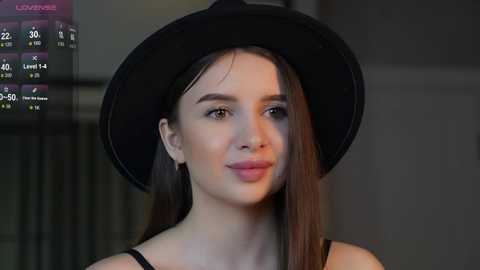 Media: Video of a young woman with fair skin, straight brown hair, wearing a black cowboy hat and black top, standing against a blurred, dimly lit background.