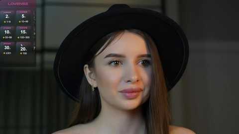 Media: Video of a young woman with light skin, brown hair, wearing a black hat and black top, with a blurred background featuring a digital display showing a weather forecast.