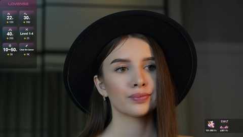 Media: Video of a young woman with fair skin and long dark hair, wearing a black wide-brimmed hat, standing indoors with a blurred, modern background.