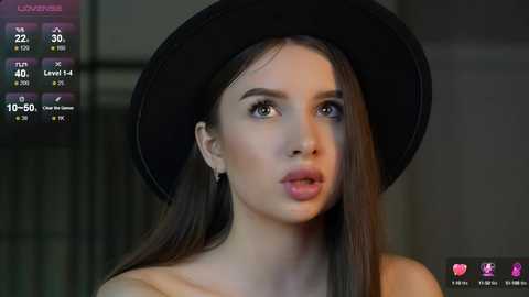 Media: Video of a fair-skinned woman with long dark hair, wearing a black hat and a black top, looking directly at the camera with a neutral expression.