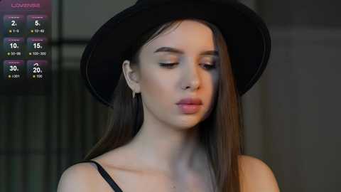 Media: Video of a young woman with long brown hair, wearing a black hat and black top, looking introspective. Background is blurred.