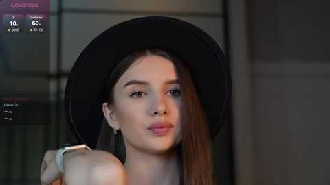 Media: Video of a fair-skinned, dark-haired woman with a large black hat, wearing a light blue top, standing indoors, blurred background.