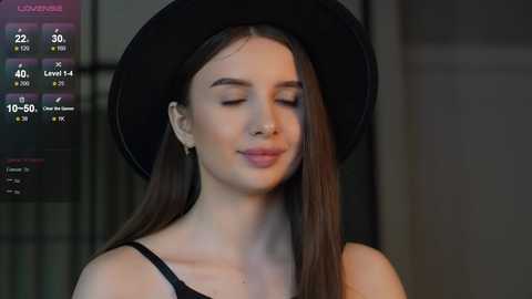 Media: Video of a serene young woman with long brown hair, wearing a black hat and black top, set against a dimly lit background.